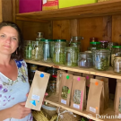Photographie couleur de l'herboriste Dorianne Herrera, debout devant une étagère sur laquelle sont présentés les produits qu'elle propose à la vente, dans des bocaux en verre et des sachets en papiers