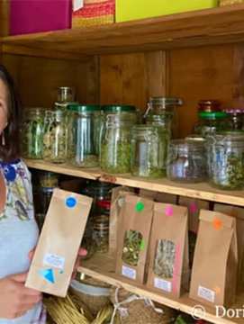 Photographie couleur de l'herboriste Dorianne Herrera, debout devant une étagère sur laquelle sont présentés les produits qu'elle propose à la vente, dans des bocaux en verre et des sachets en papiers