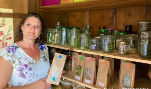Photographie couleur de l'herboriste Dorianne Herrera, debout devant une étagère sur laquelle sont présentés les produits qu'elle propose à la vente, dans des bocaux en verre et des sachets en papiers