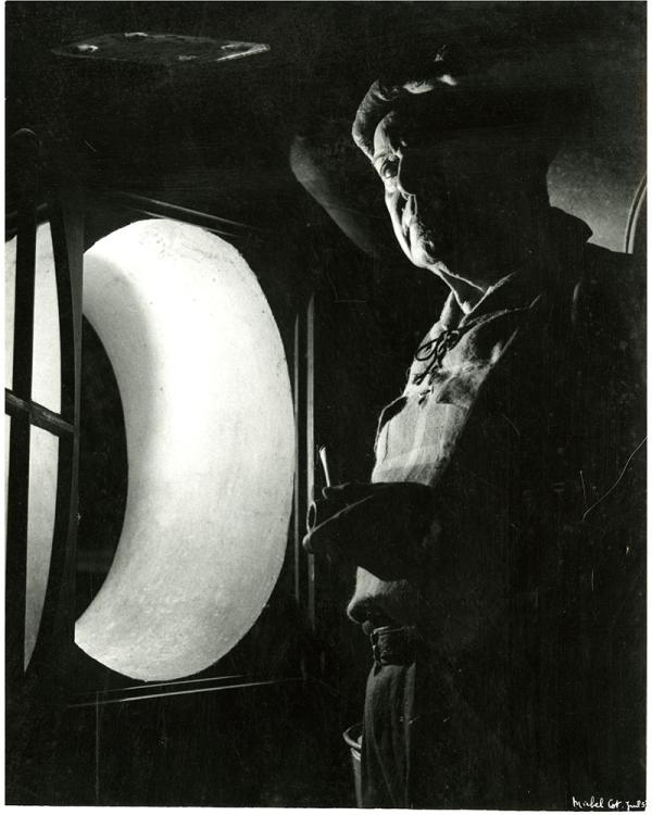 Portrait en noir et blanc de Pierre Mac Orlan. Il se tient dans l'obscurité devant la fenêtre dite "œil de bœuf" dans sa maison